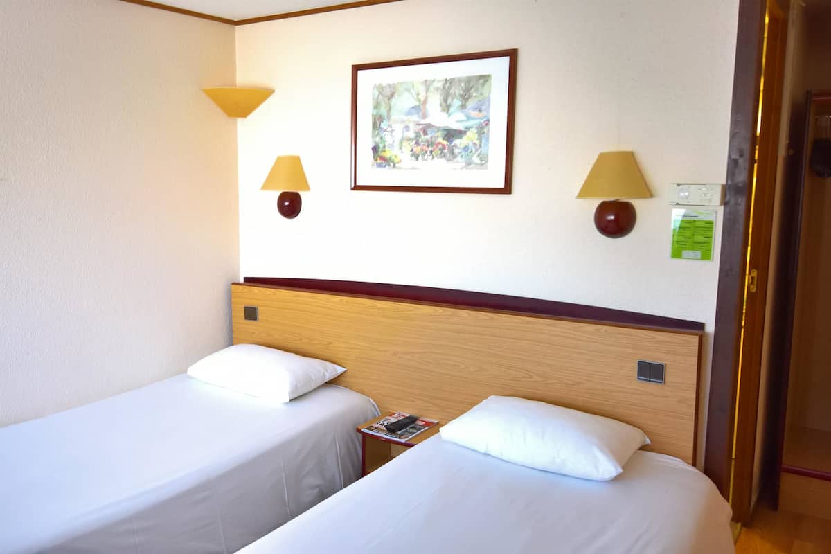 Una habitación de hotel con dos camas individuales, un cabecero de madera, un cuadro enmarcado y dos lámparas de pared.