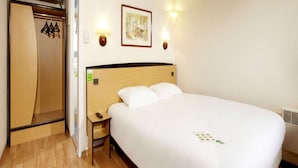 Une chambre d’hôtel avec une tête de lit en bois, un lit simple, une table de chevet, une petite table et une armoire avec des cintres.