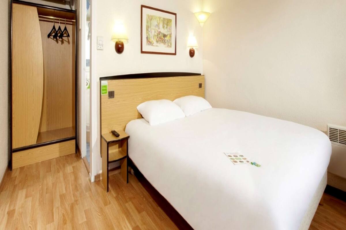 Habitación de hotel con cabecera de madera, una cama individual, una mesita de noche, una pequeña mesa y un armario con perchas.