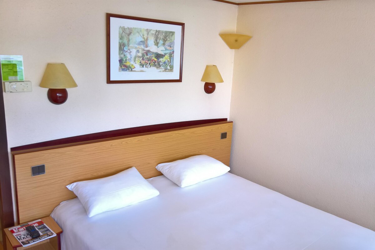 A hotel room with a wooden headboard, two white pillows, a framed painting on the wall, and two wall-mounted lamps.