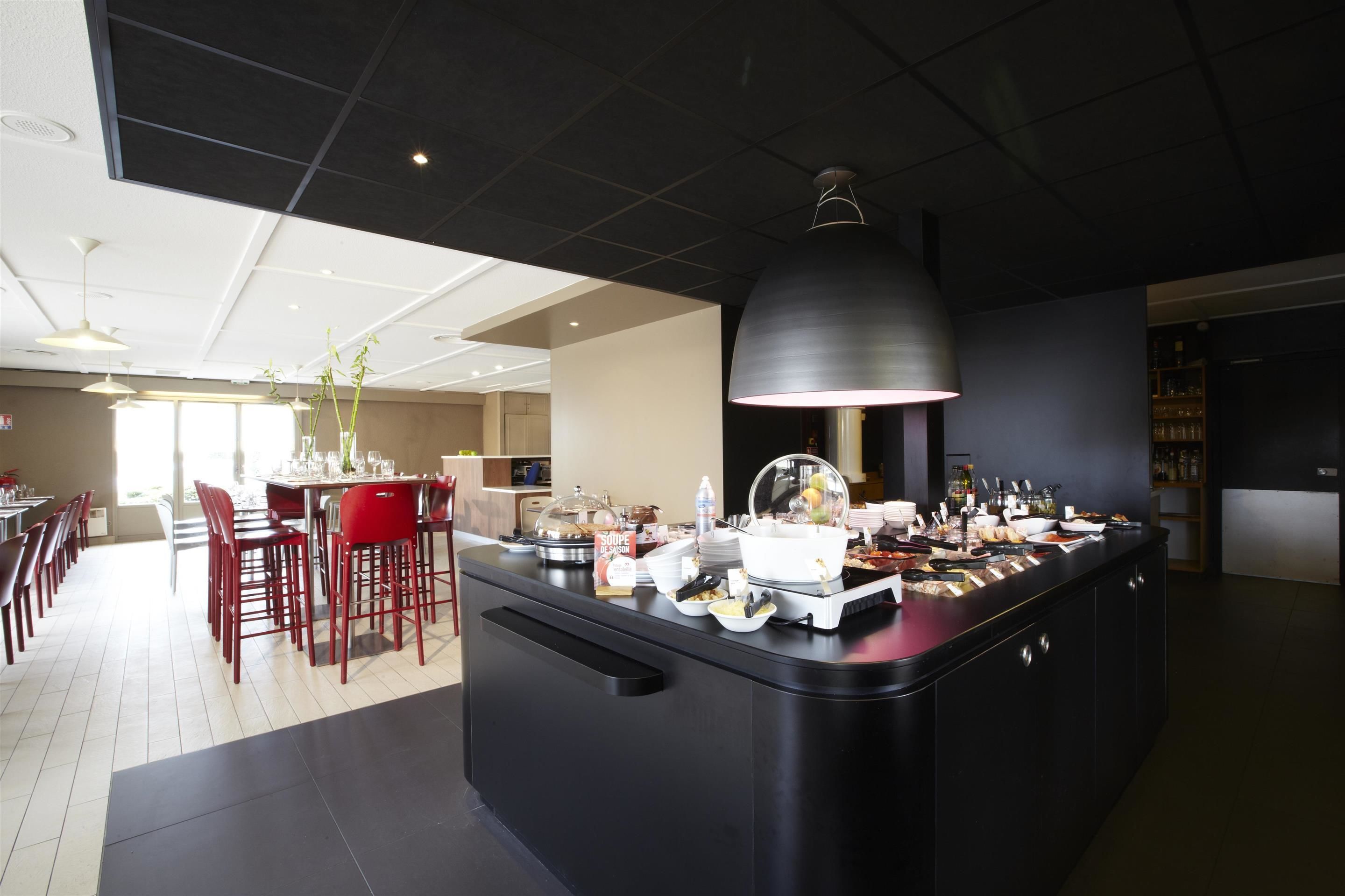 A modern hotel lobby with a central buffet area, red chairs, and a large pendant light.