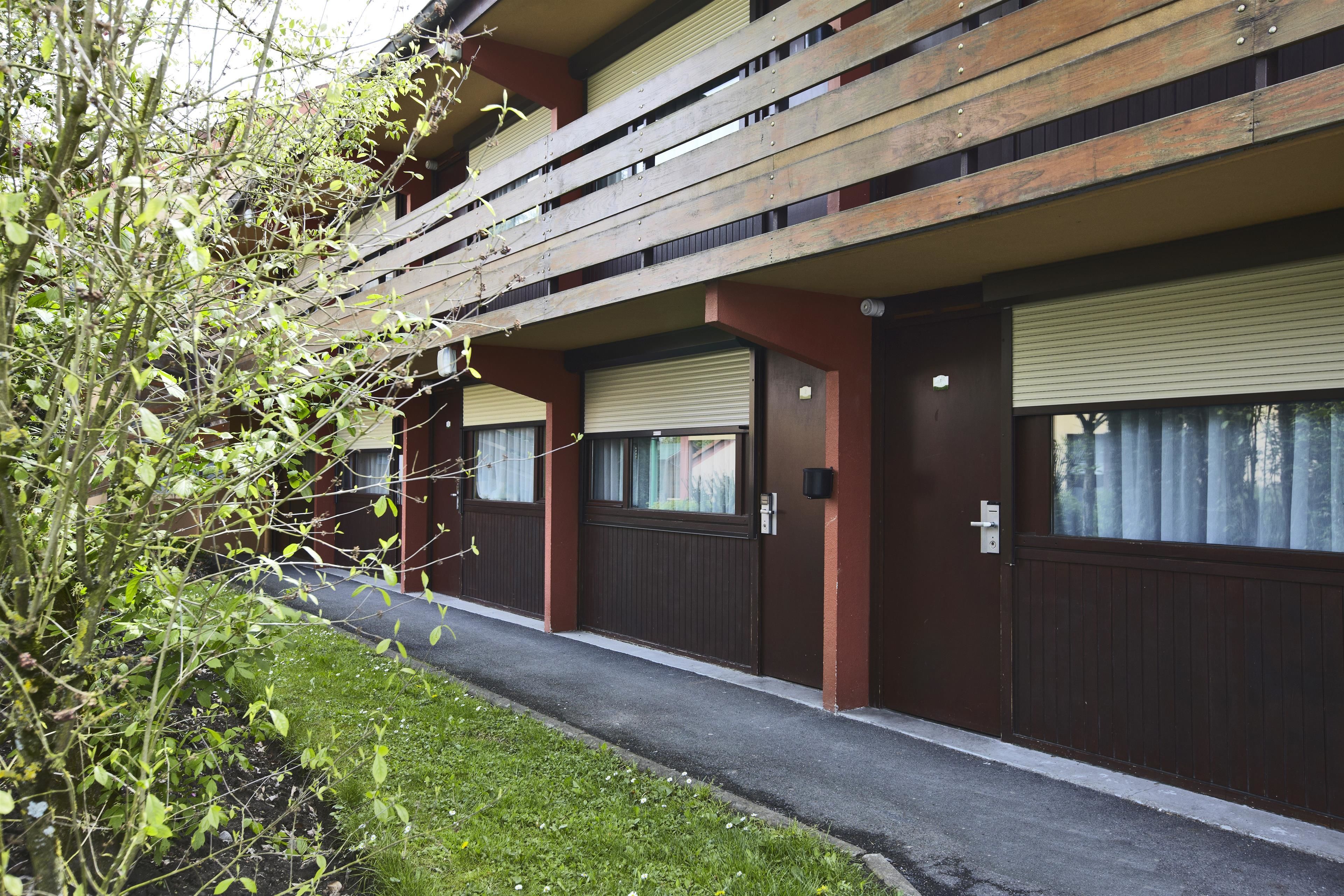 A row of residential-style buildings with wooden balconies, a paved walkway, and a grassy area.