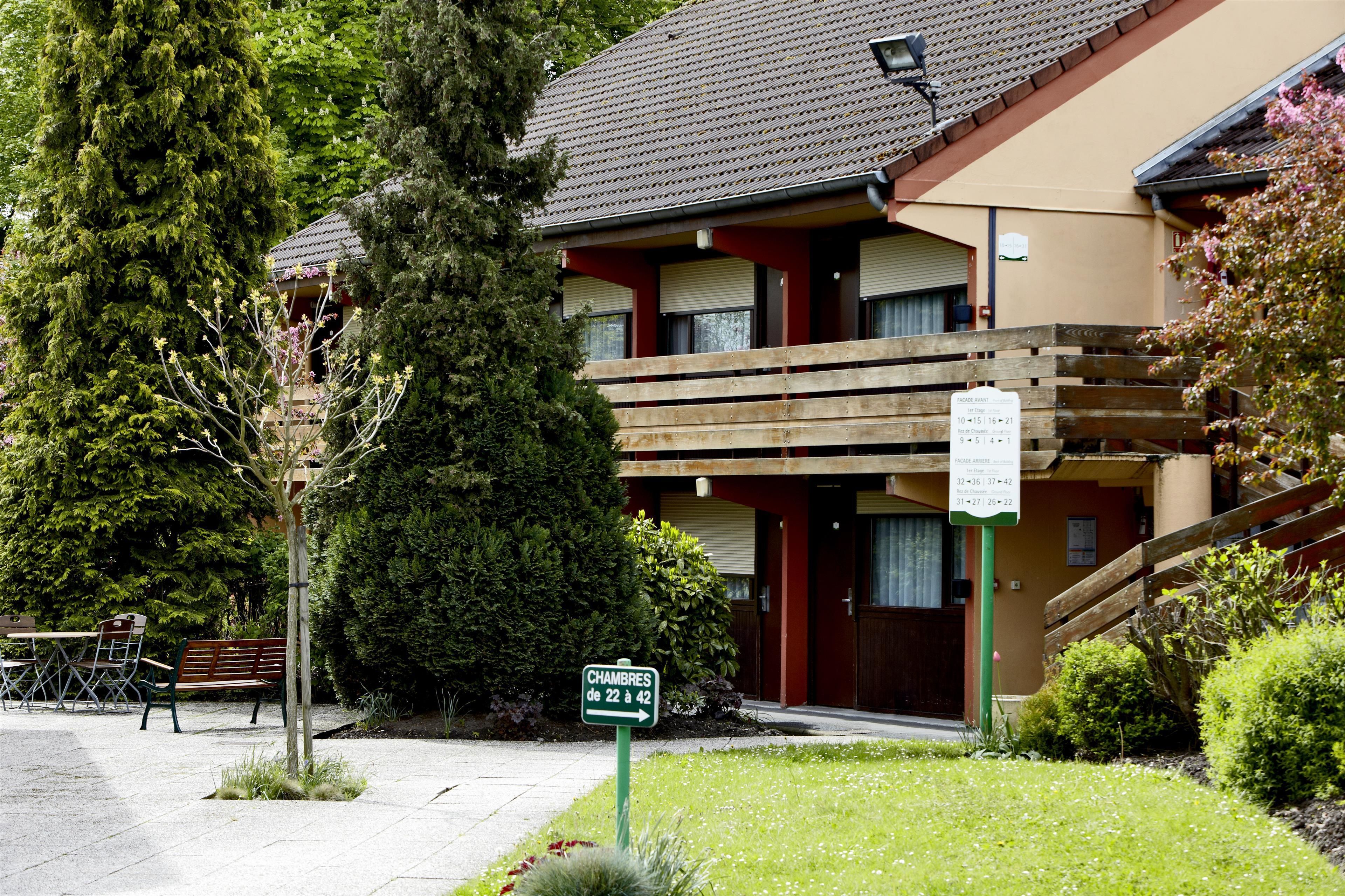 Un edificio de dos pisos con balcón de madera, un letrero que indica los números de las habitaciones y un jardín bien cuidado.