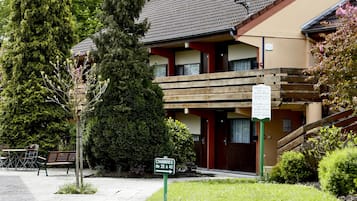 Un edificio de dos pisos con balcón de madera, un letrero que indica los números de las habitaciones y un jardín bien cuidado.
