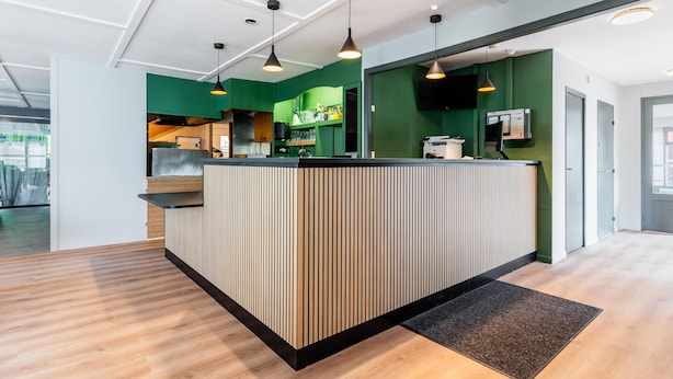 A modern reception area with a wooden reception desk, green walls, and pendant lighting. - Campanile NATURE - Thionville Yutz (Yutz)