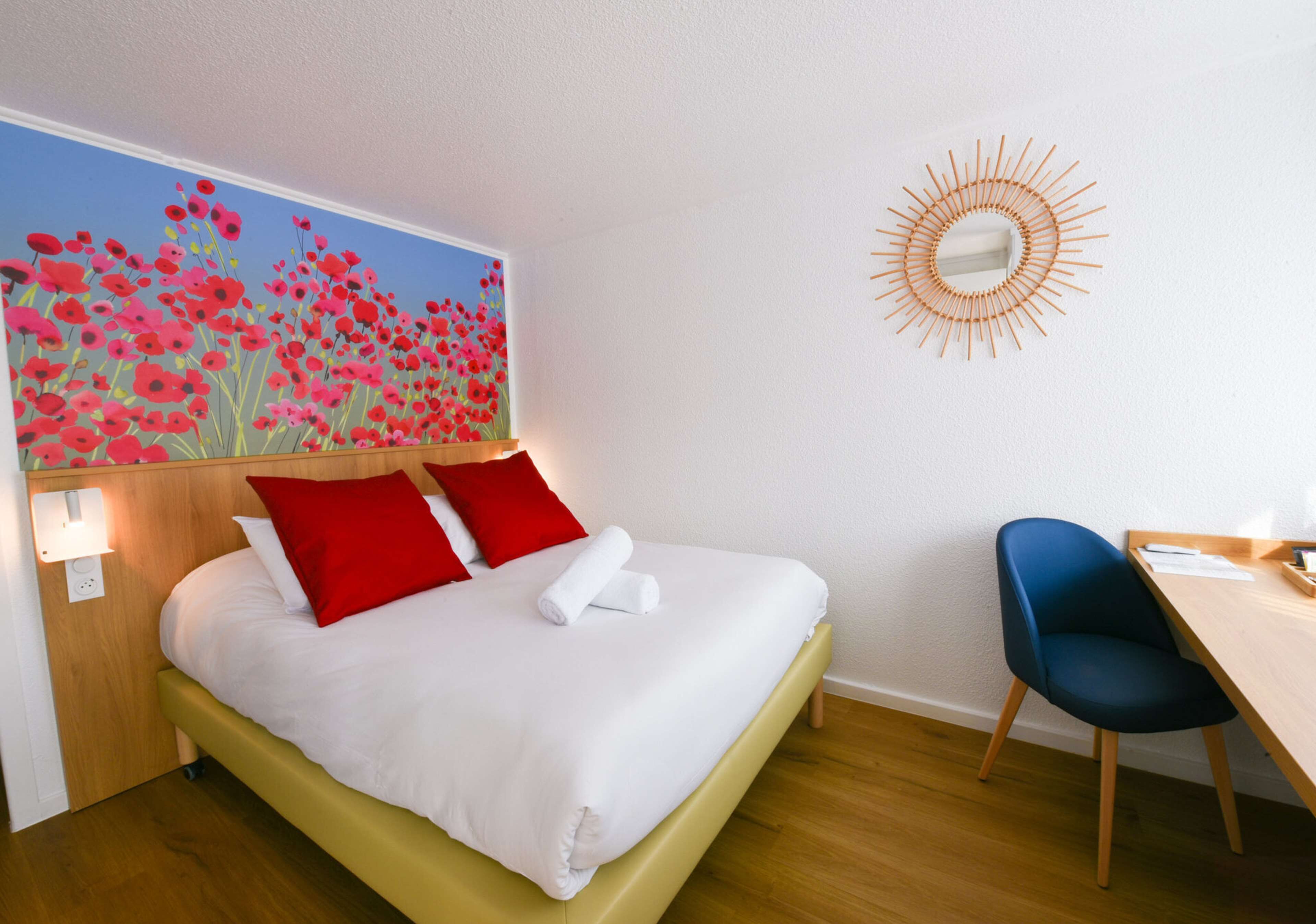 A hotel room with a bed, a desk, a chair, a wall painting, and a sunburst light fixture.
