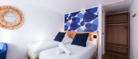 A hotel room with two beds, a wooden wardrobe, a round mirror, and a wall with a blue abstract design.