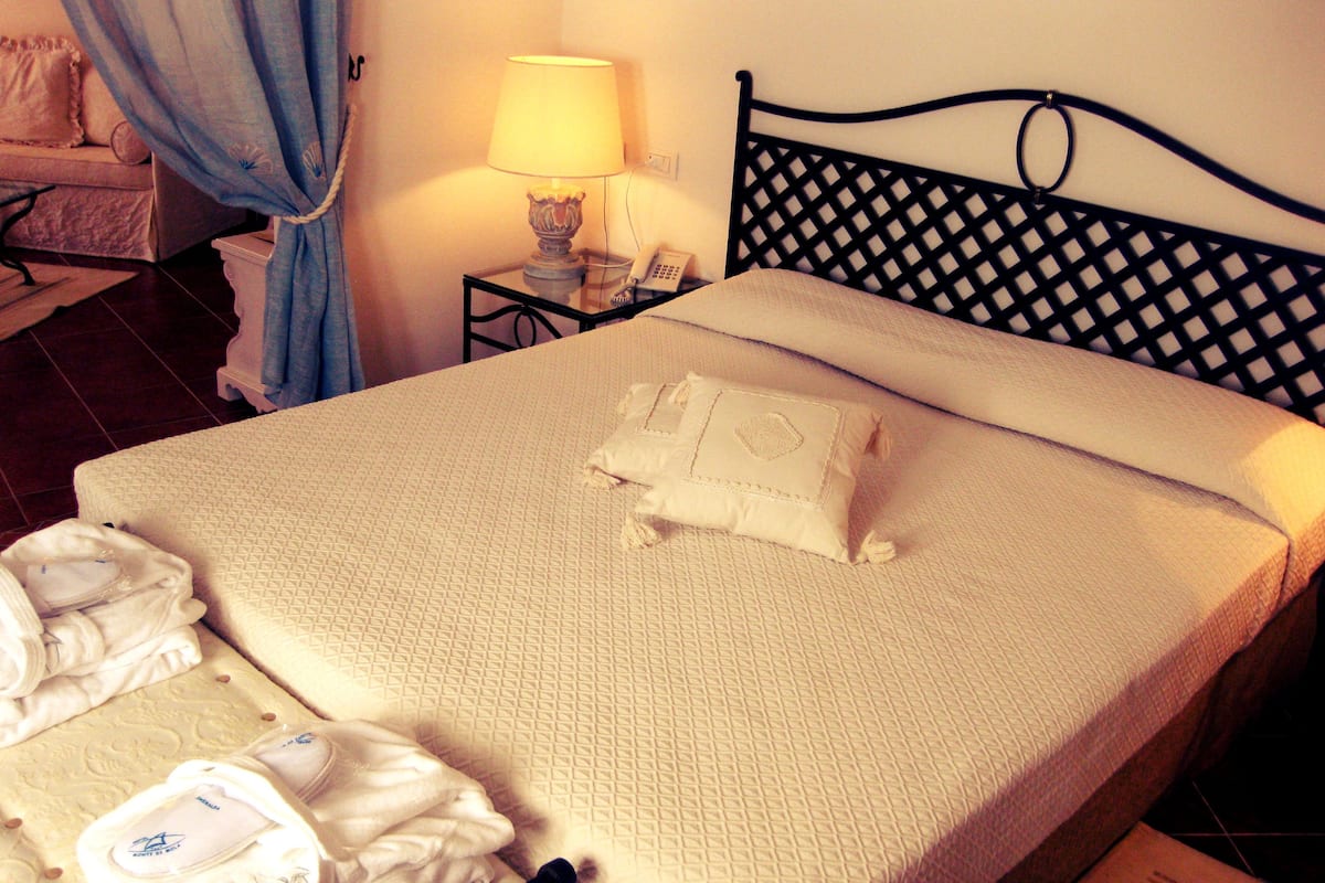 Un lit bien fait, avec une couette blanche et un coussin décoratif, une table de chevet avec une lampe, et un rideau bleu partiellement tiré.