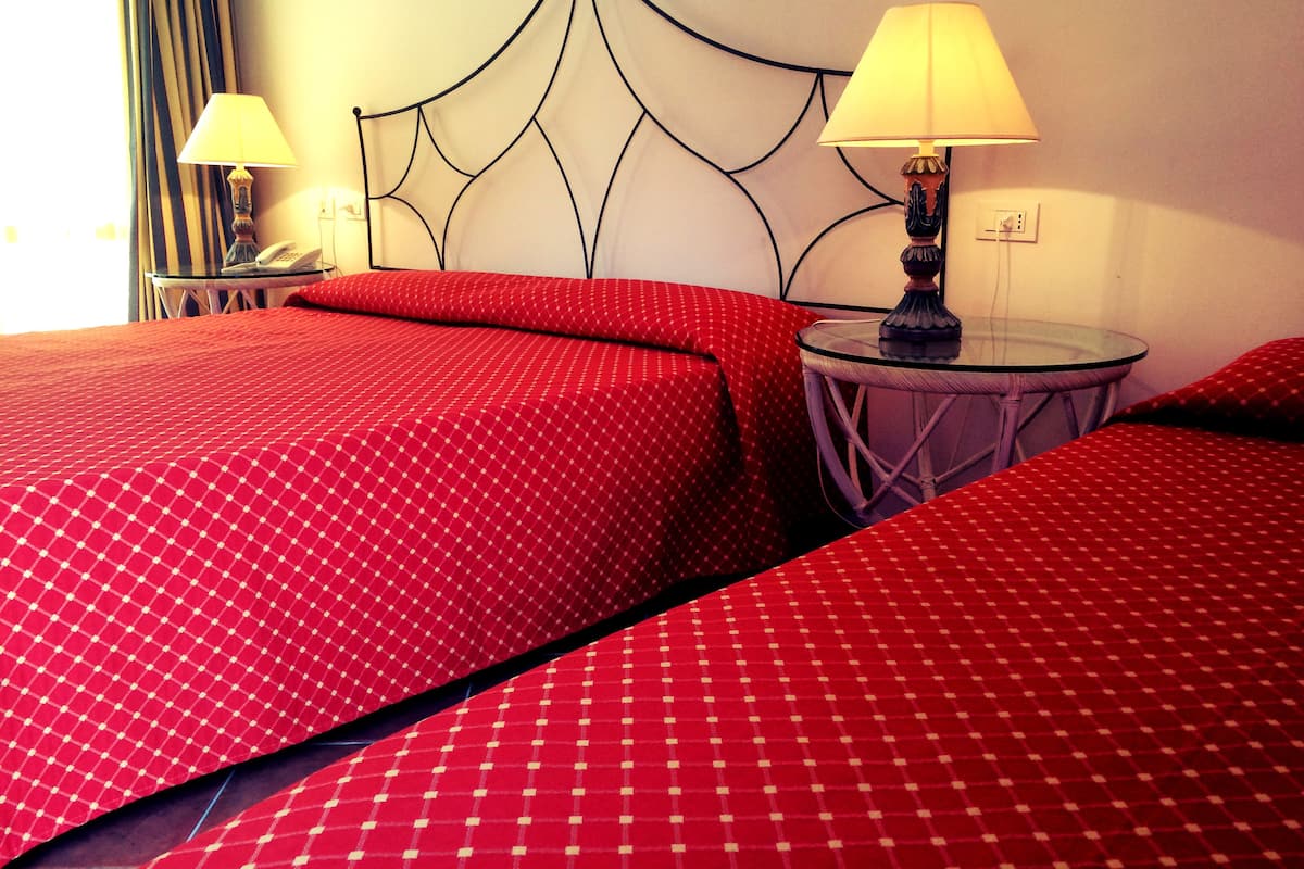 Une chambre d’hôtel avec deux lits, chacun agrémenté de draps rouges et d’une tête de lit décorative. On y trouve deux tables de chevet avec des lampes, ainsi qu’une petite table également dotée d’une lampe.