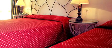 Una habitación de hotel con dos camas, cada una con ropa de cama roja y un cabecero decorativo. Hay dos mesitas de noche con lámparas y una pequeña mesa con una lámpara.