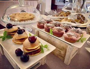 Een buffet met diverse gebakjes en desserts, waaronder pannenkoeken met kersen, muffins en een taart met een deksel.
