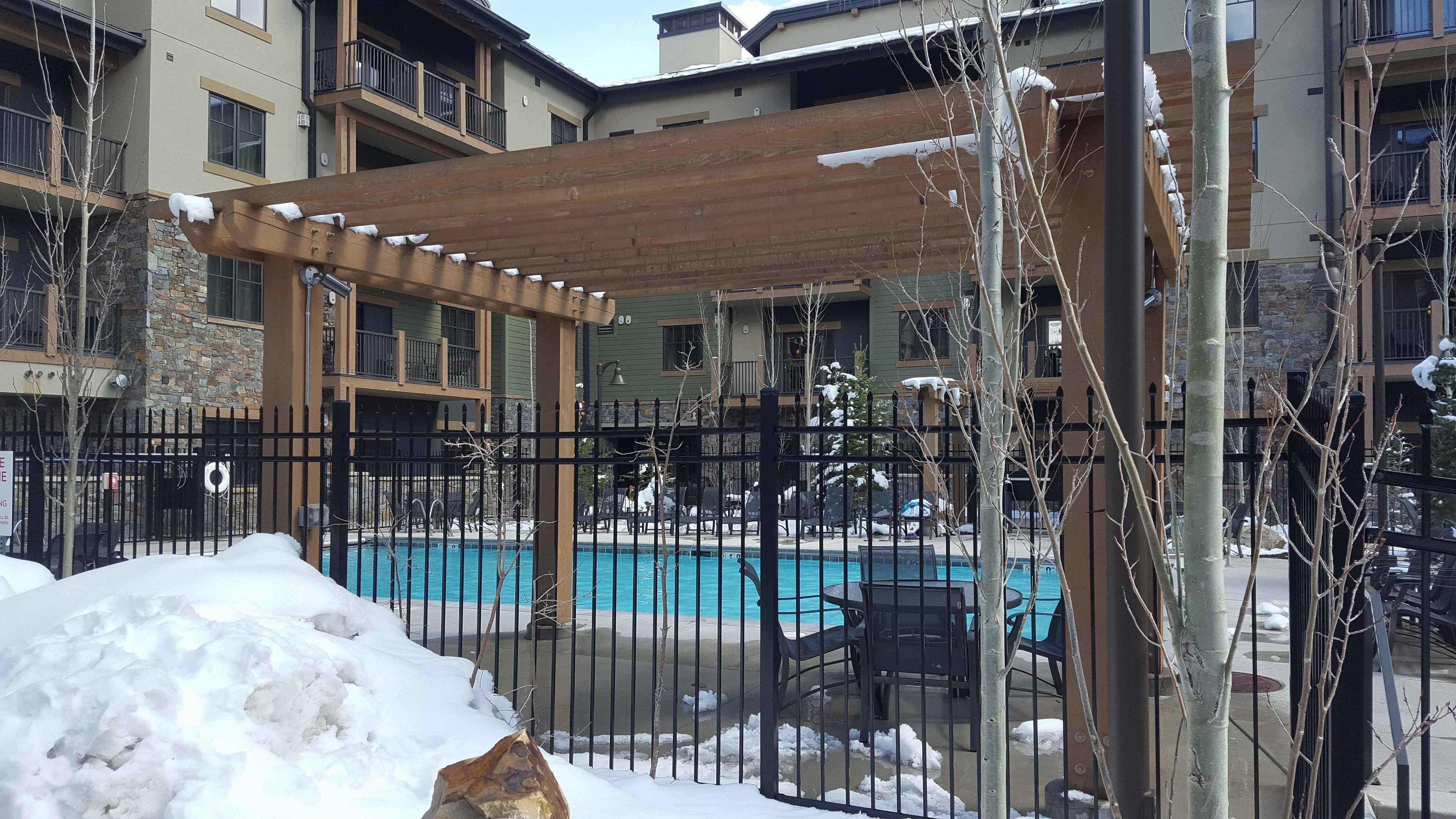 A pool area with a wooden pergola, a fenced patio, and a snow-covered ground.