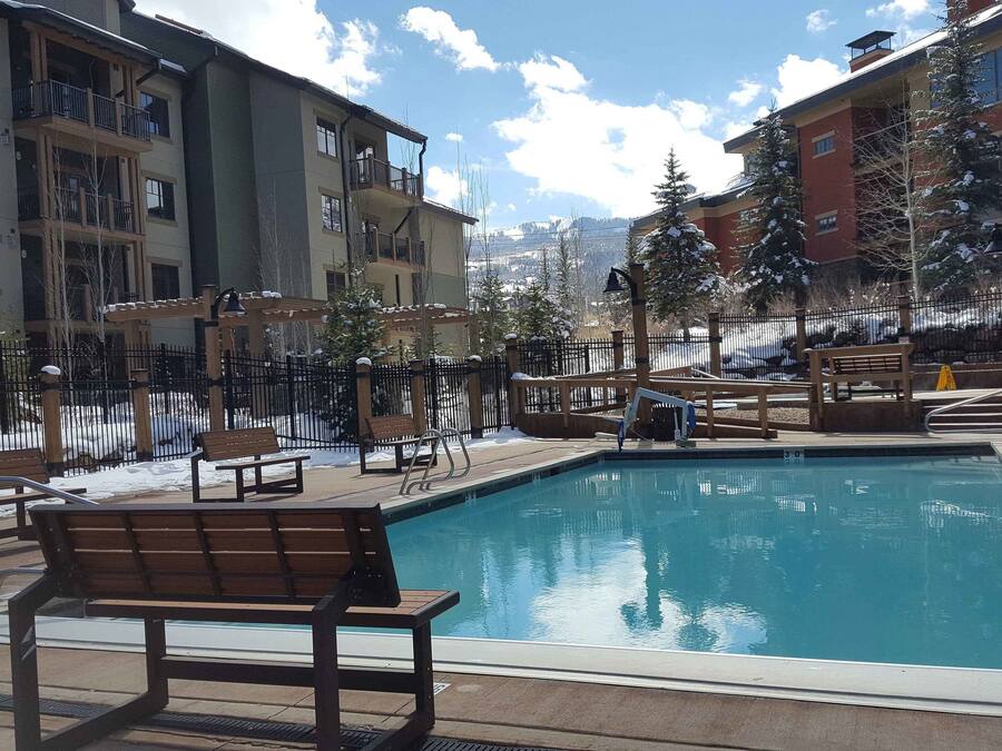 En swimmingpool med en träbänk i förgrunden, omgiven av byggnader och snötäckta träd.