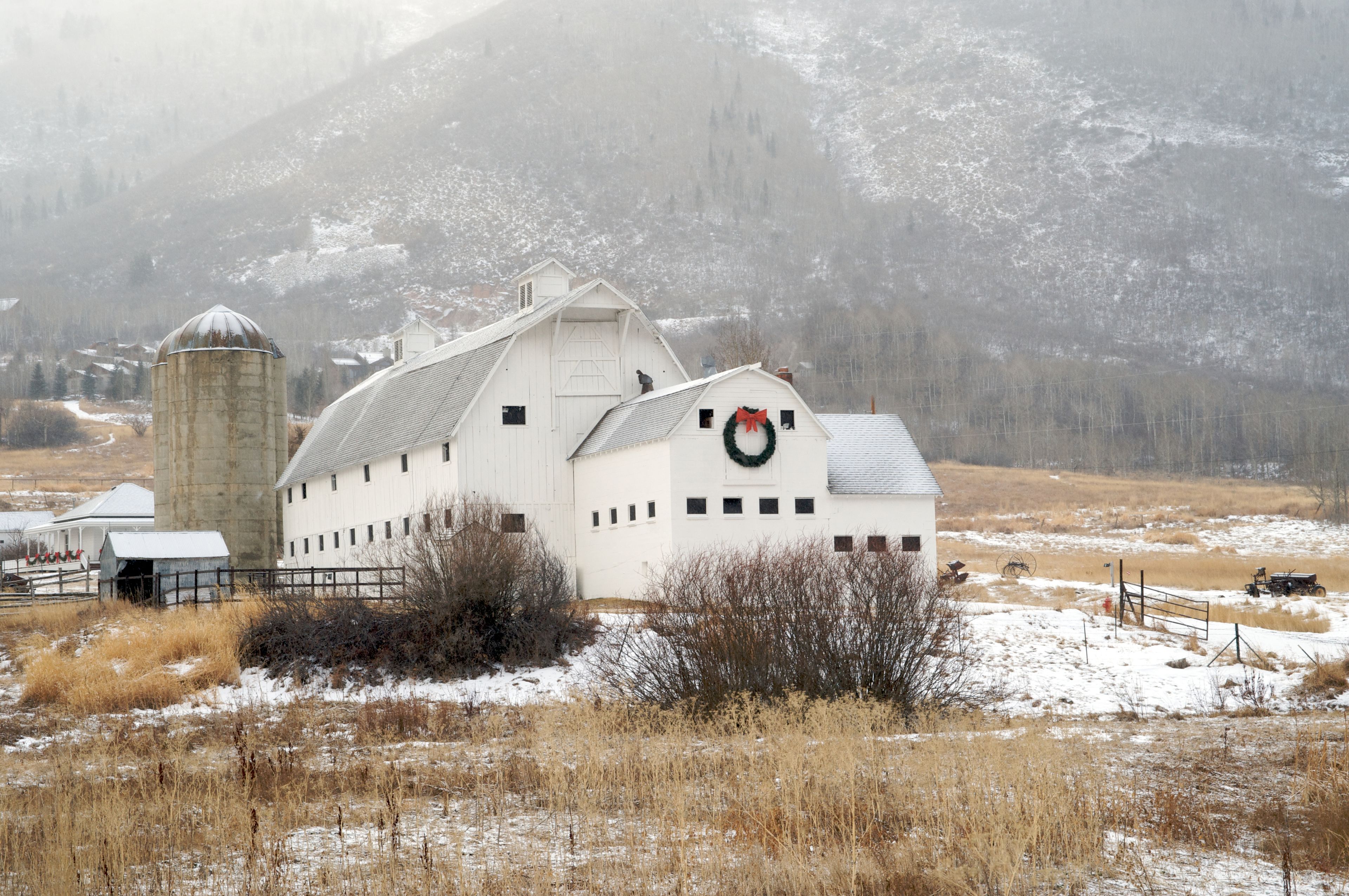 A white barn with a green wreath on the door, a silo, and a tractor in a snowy field.
