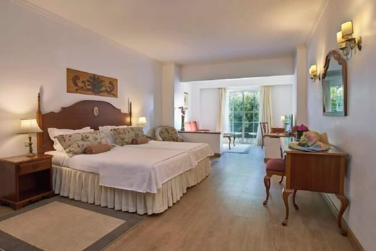 Ein Hotelzimmer mit Doppelbett, Schreibtisch, Stuhl, Lampe und einem Fenster mit Blick auf Grünflächen.