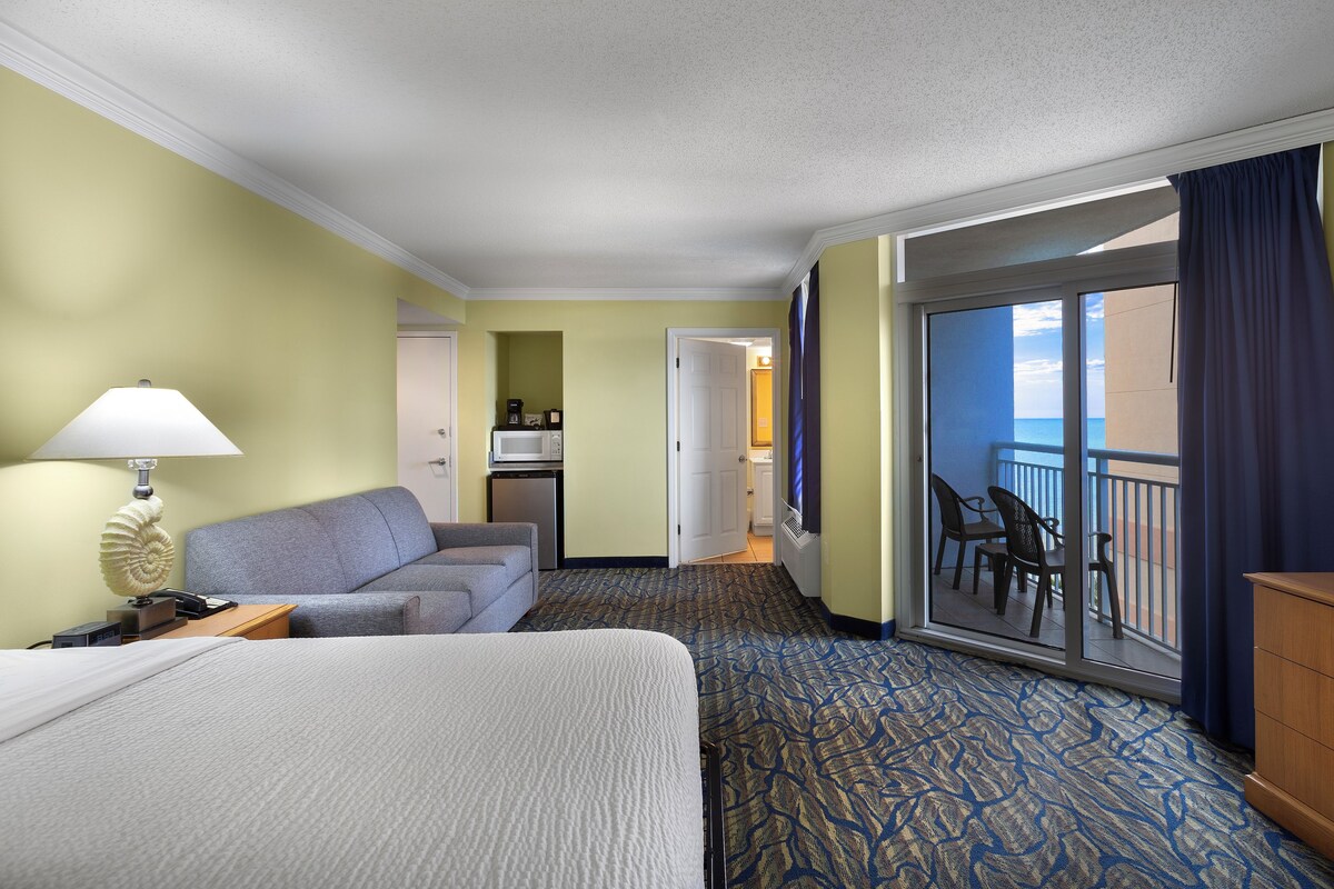 A hotel room with a bed, a sofa, a desk with a lamp, a dresser, and a balcony with a view of the ocean.
