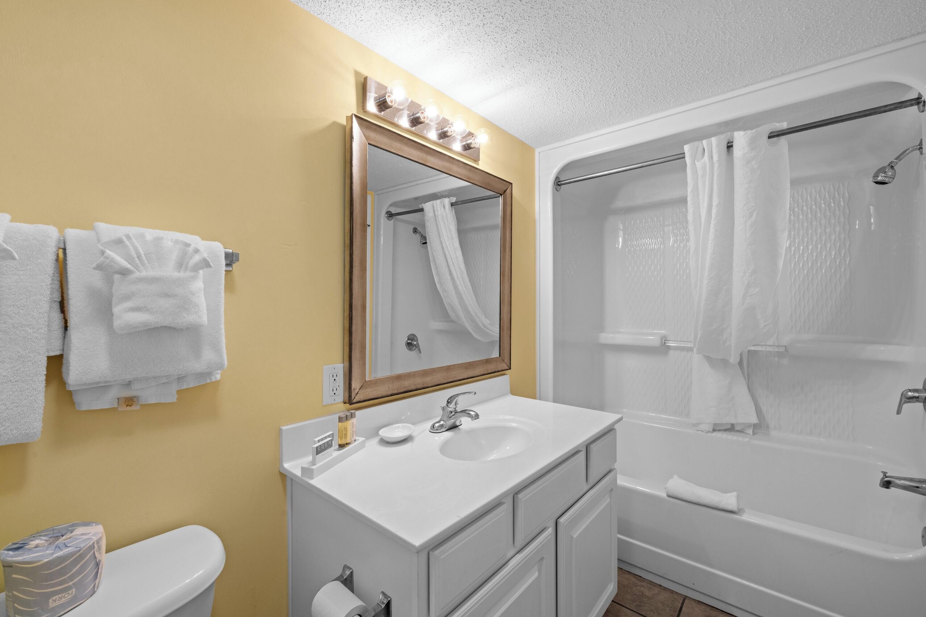 A bathroom with a white sink, mirror, and bathtub. There are towels hanging on the wall and a toilet paper roll on the counter.