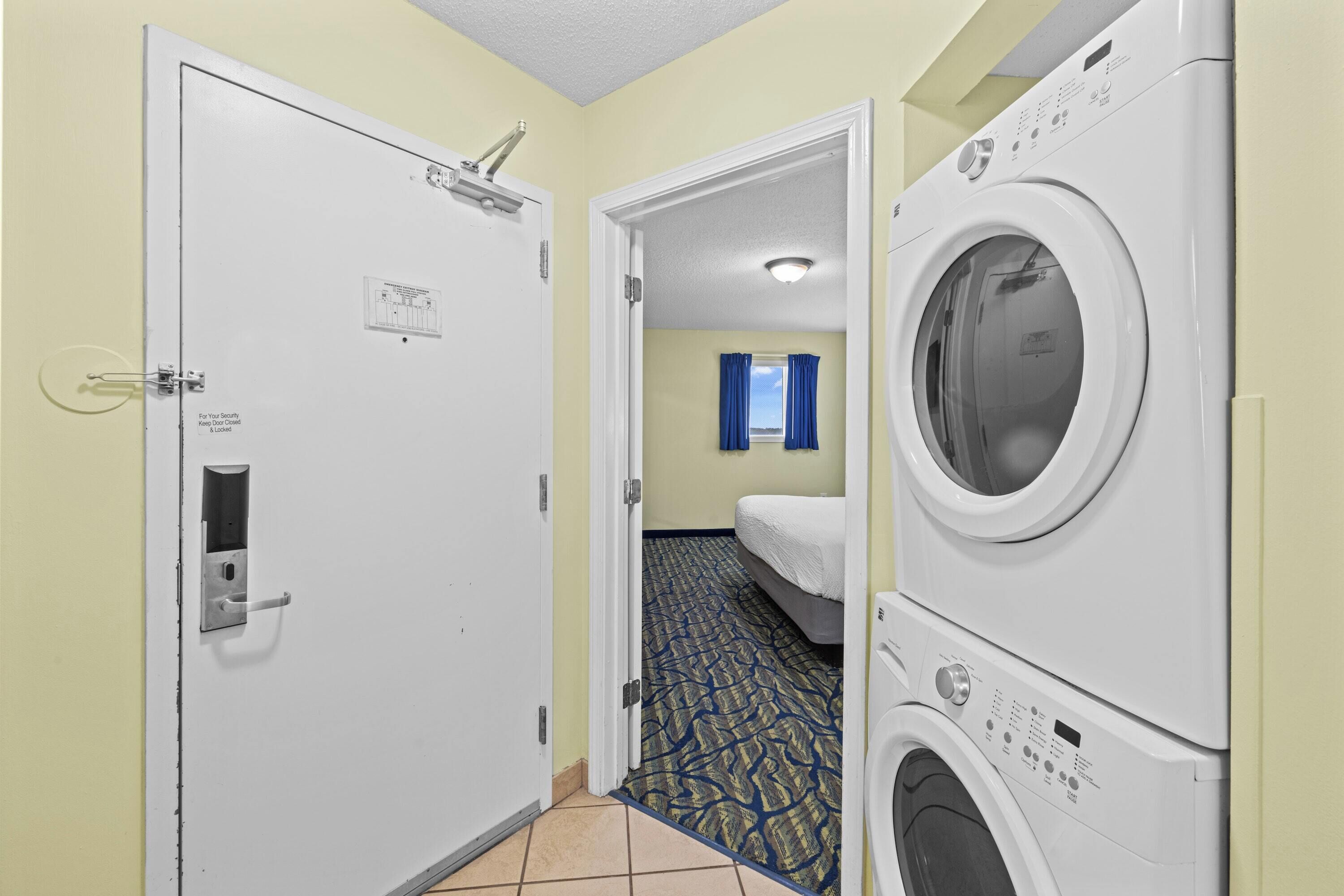 A hotel room with a door, a washing machine, a bed, and a patterned carpet.