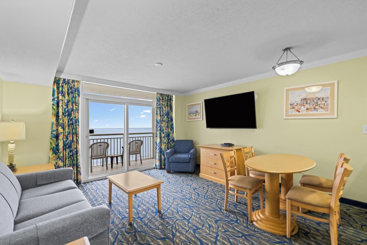 A hotel room with a sofa, a dining table with chairs, a TV mounted on the wall, and a balcony with a view.
