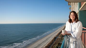 A person in a white robe holding a cup, standing on a balcony overlooking a beach with waves and a clear sky.