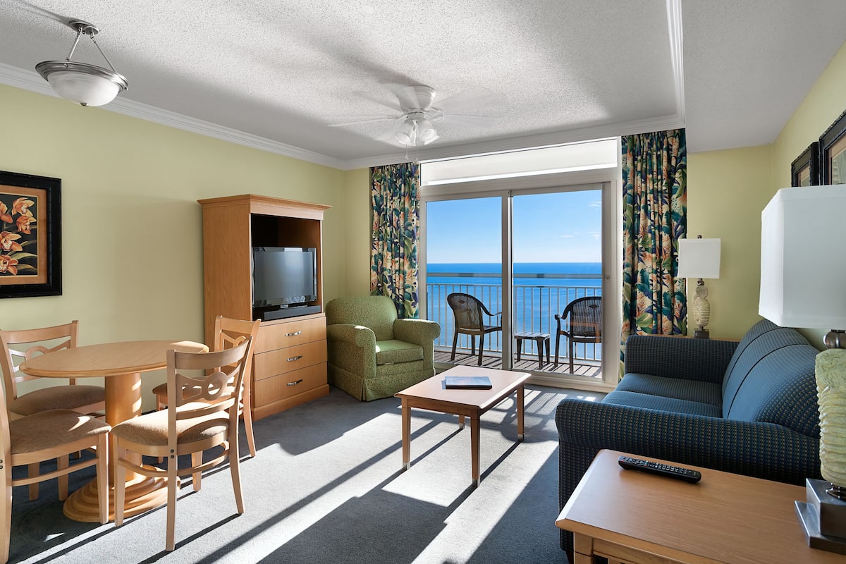A hotel room with a dining area, a sofa, a TV, a balcony with a view of the sea, and a ceiling fan.