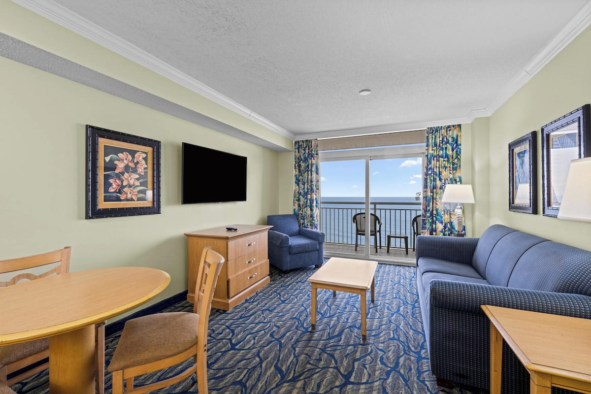 A hotel room with a balcony, a sofa, a dining table, and a TV.