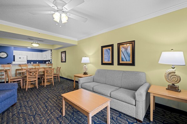 A hotel room with a gray sofa, a wooden coffee table, a blue armchair, a dining area with a table and chairs, a kitchen area, and a ceiling fan.