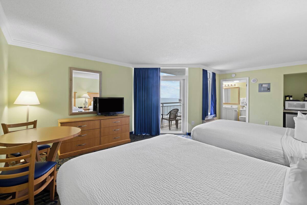 A hotel room with two beds, a desk with a chair, a dresser, a television, and a balcony with a chair.
