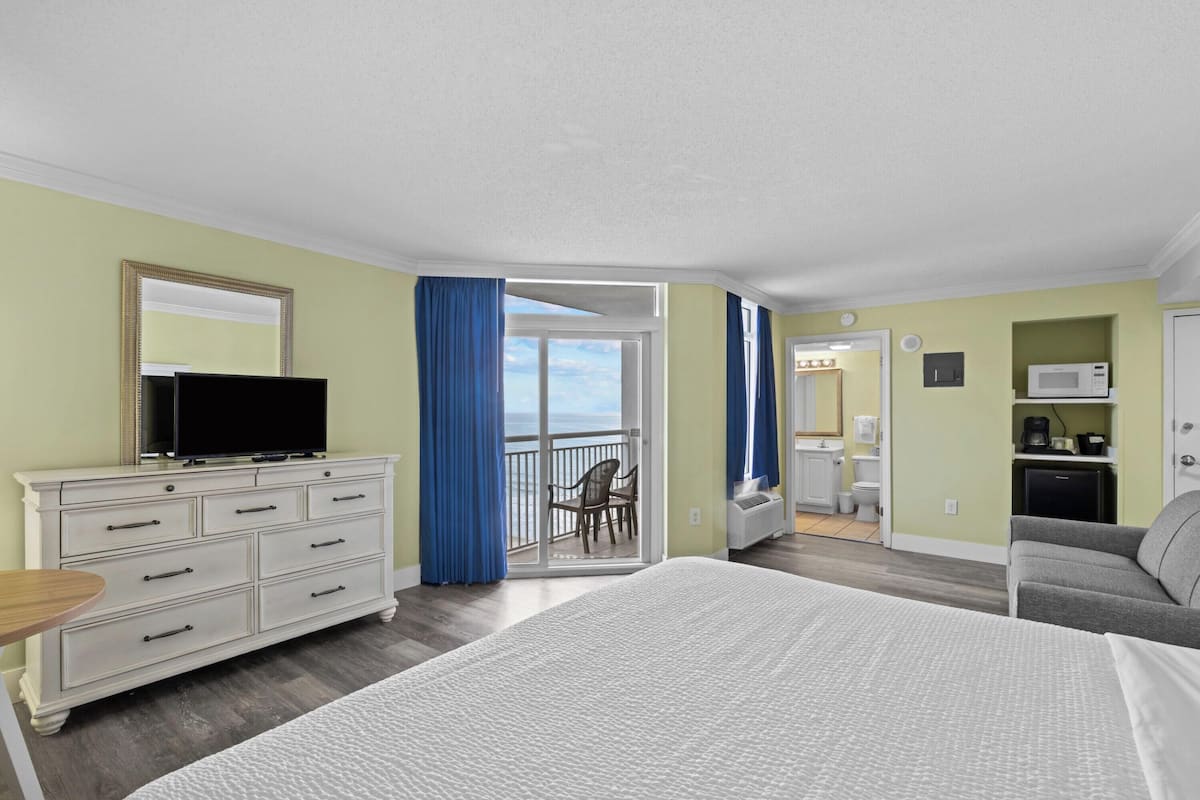 A hotel room with a bed, a TV on a dresser, a balcony with a view, and a kitchenette.