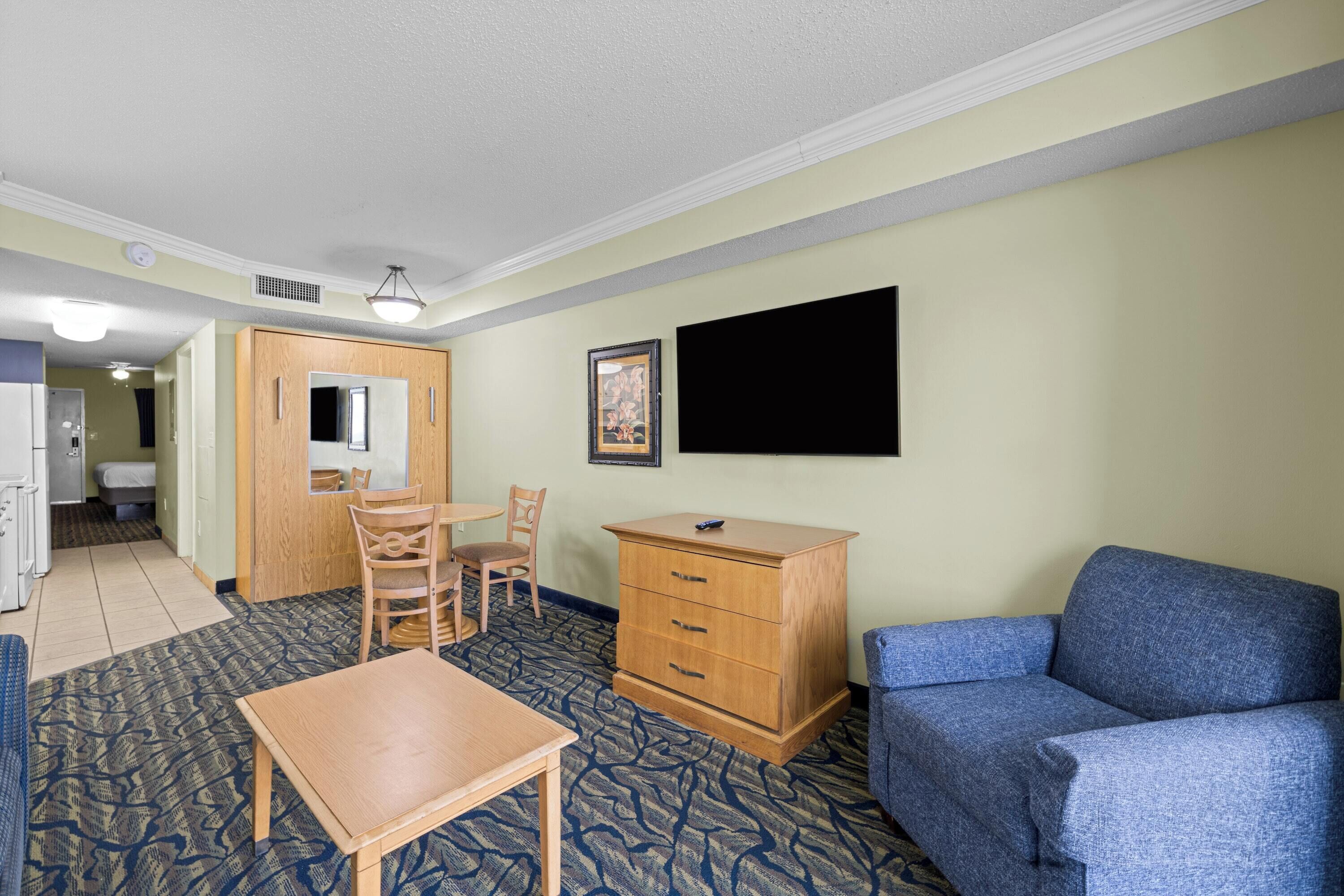 A hotel room with a dining table, chairs, a blue sofa, a wooden dresser, and a flat-screen TV mounted on the wall.