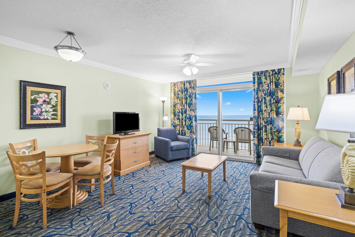 A hotel room with a balcony, a television, a sofa, a dining table with chairs, and a patterned carpet.