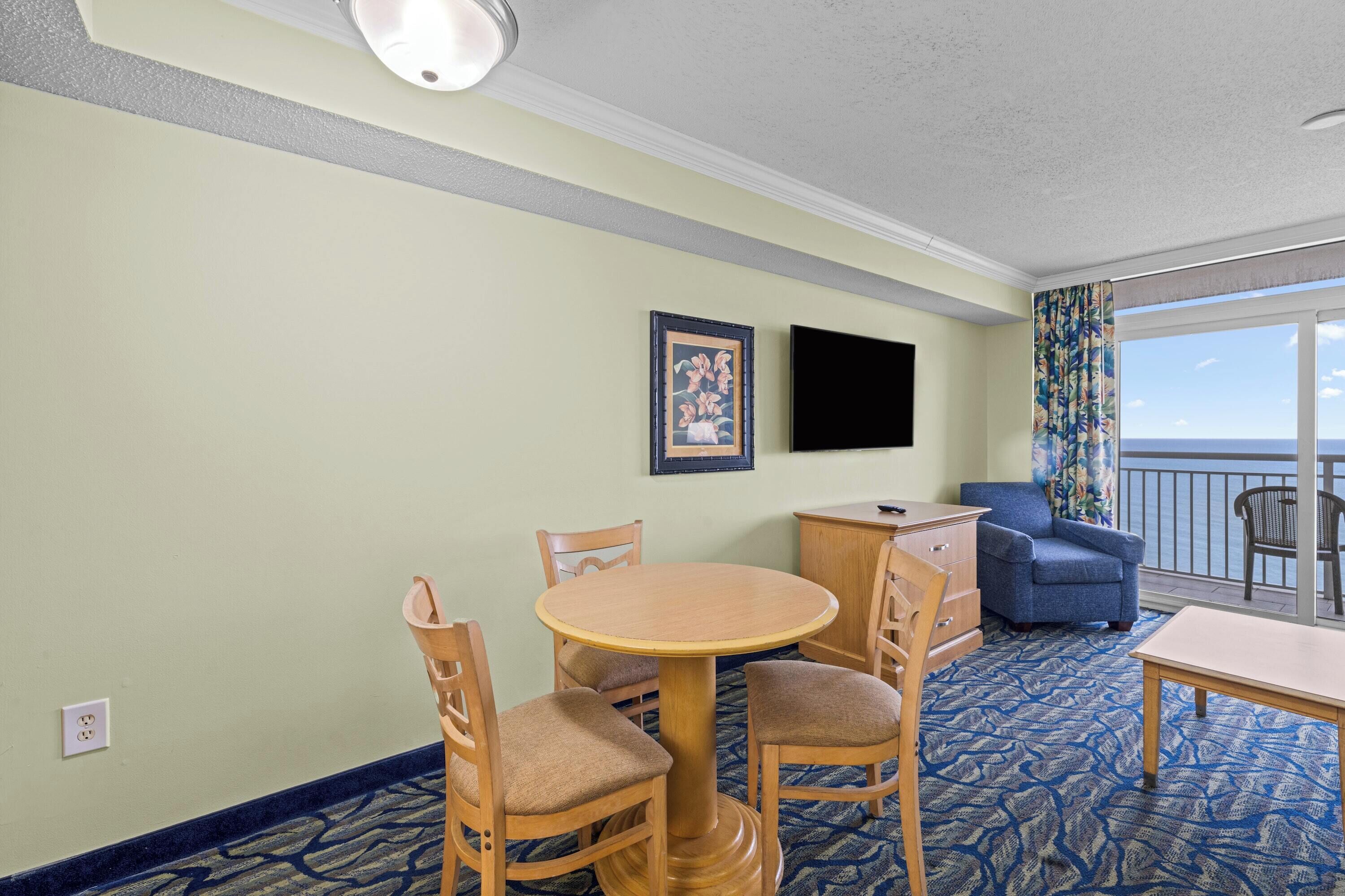 A room with a round wooden table, two chairs, a blue armchair, a TV on the wall, and a balcony with a view.