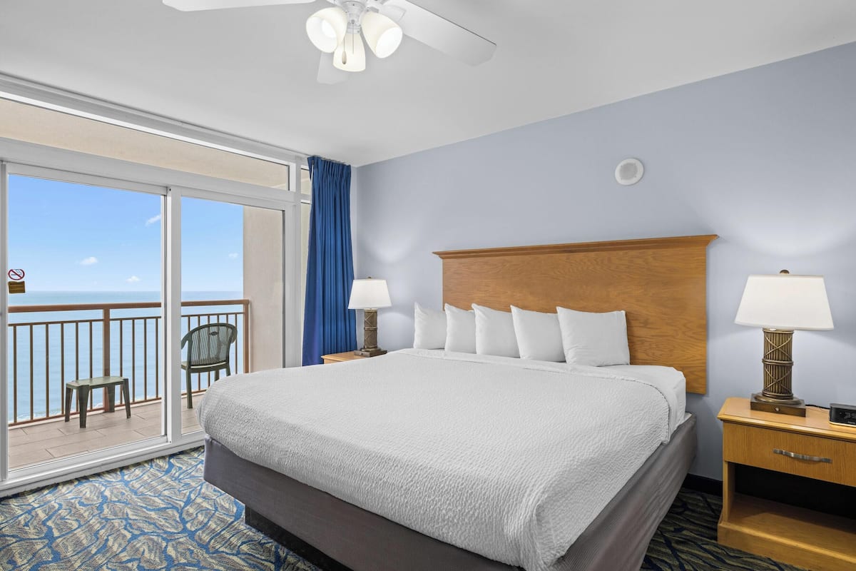 A hotel room with a large bed, two bedside tables, a ceiling fan, a view of the ocean, and a balcony with a chair.