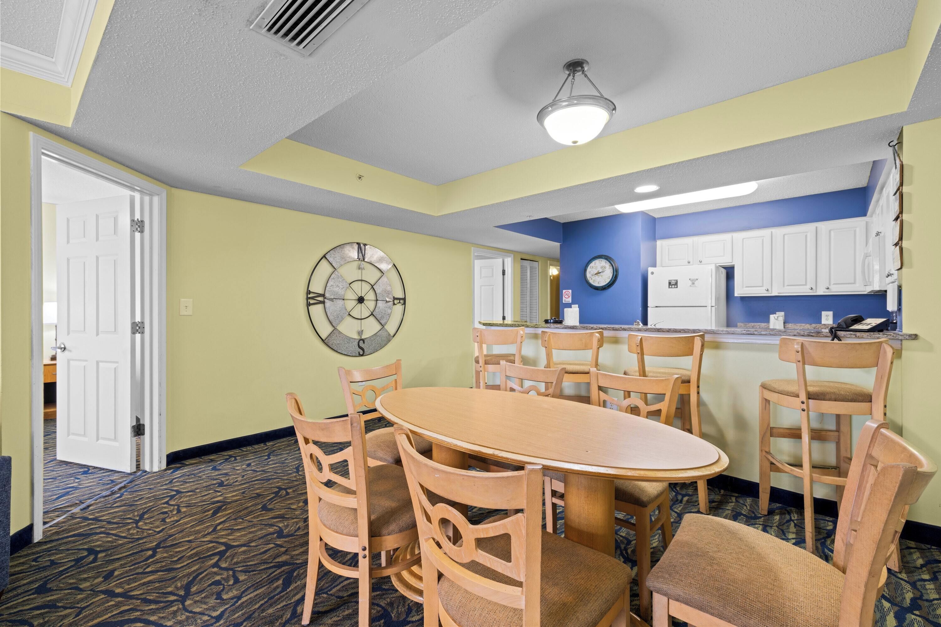 A hotel room with a dining area, a clock, and a kitchen counter.