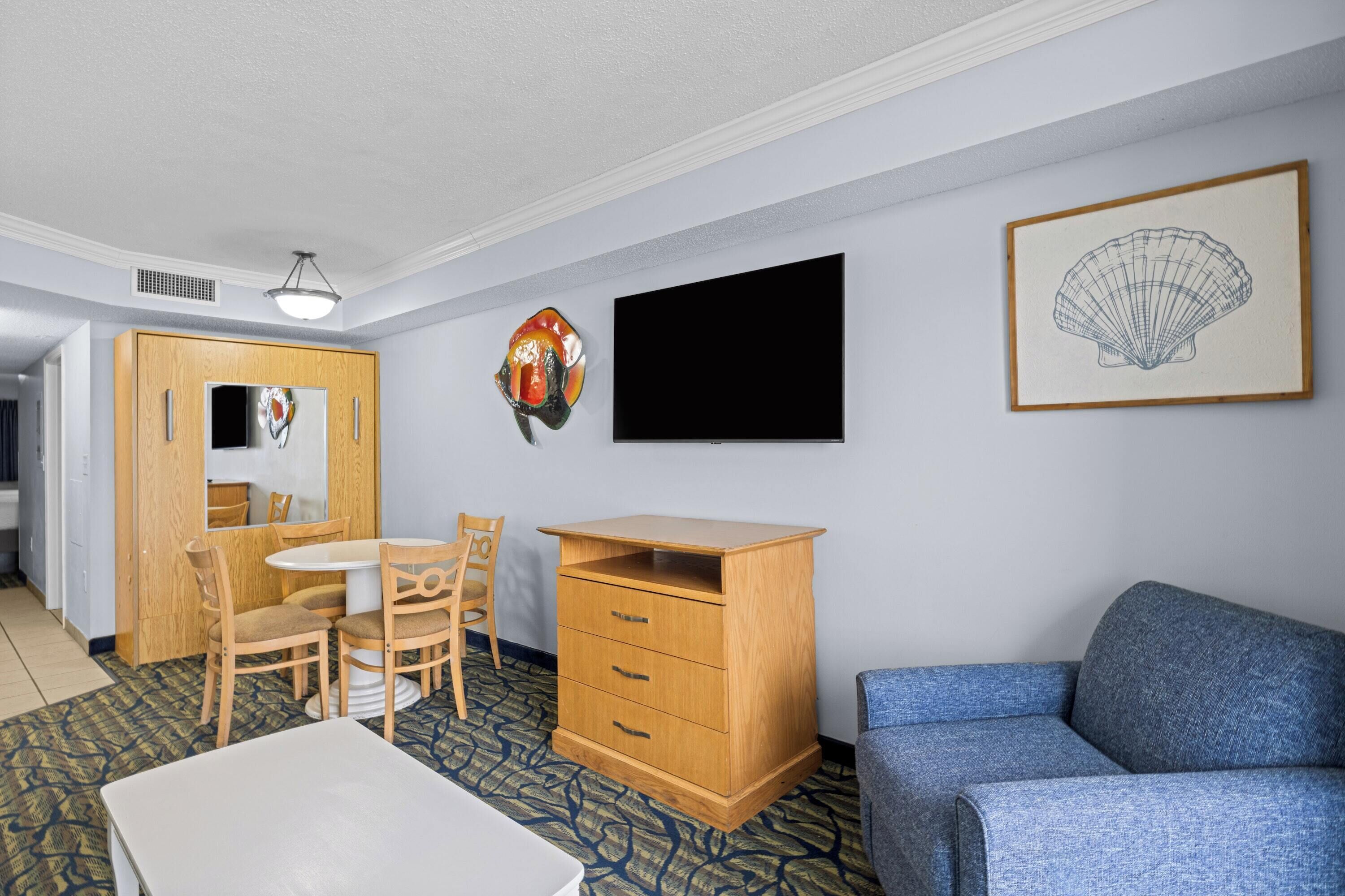 A hotel room with a dining area, a sofa, a TV, and a framed artwork on the wall.