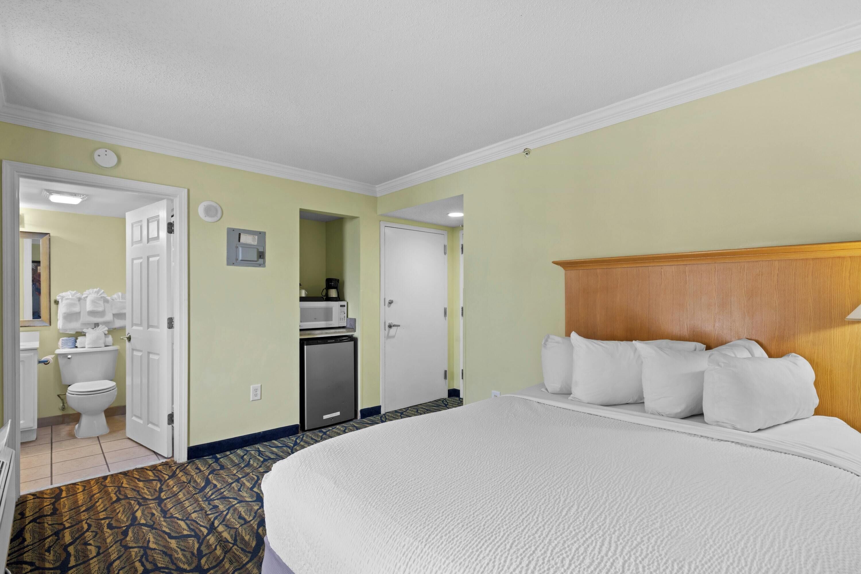 A hotel room with a large bed, a wooden headboard, a microwave, a refrigerator, and a bathroom with a toilet and sink.