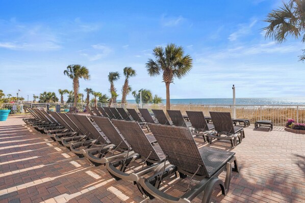 A row of wicker lounge chairs on a brick patio with a view of the ocean and palm trees.