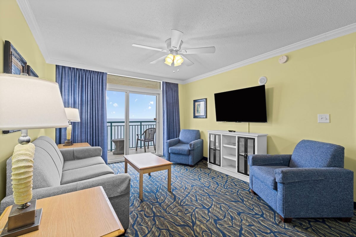 A hotel room with a ceiling fan, a flat-screen TV, a sofa, a chair, a small table, and a view of the outdoors.