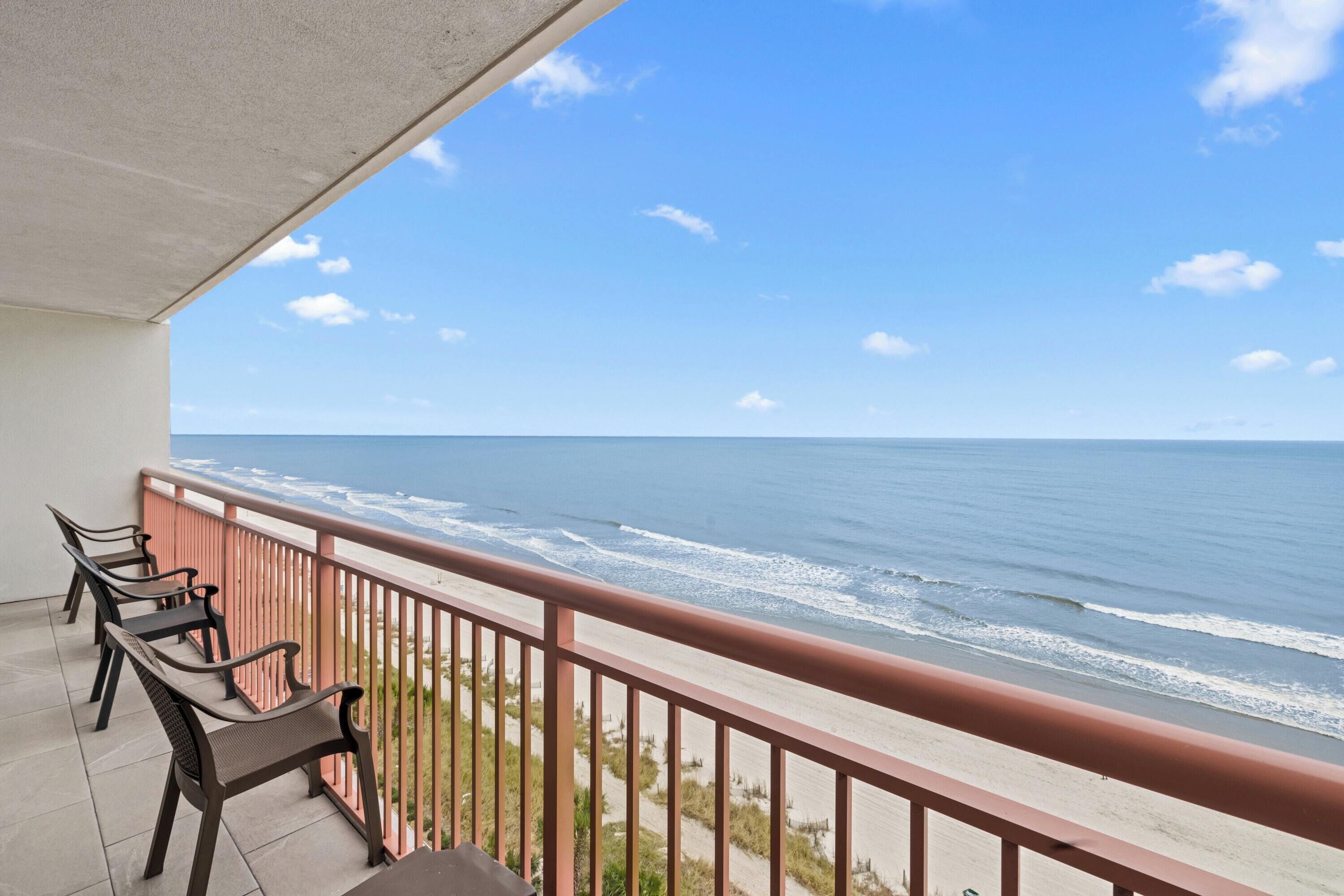 A balcony with a view of the ocean, a railing, and a row of chairs.