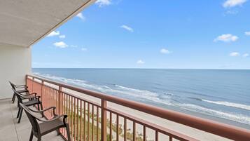 A balcony with a view of the ocean, a railing, and a row of chairs.