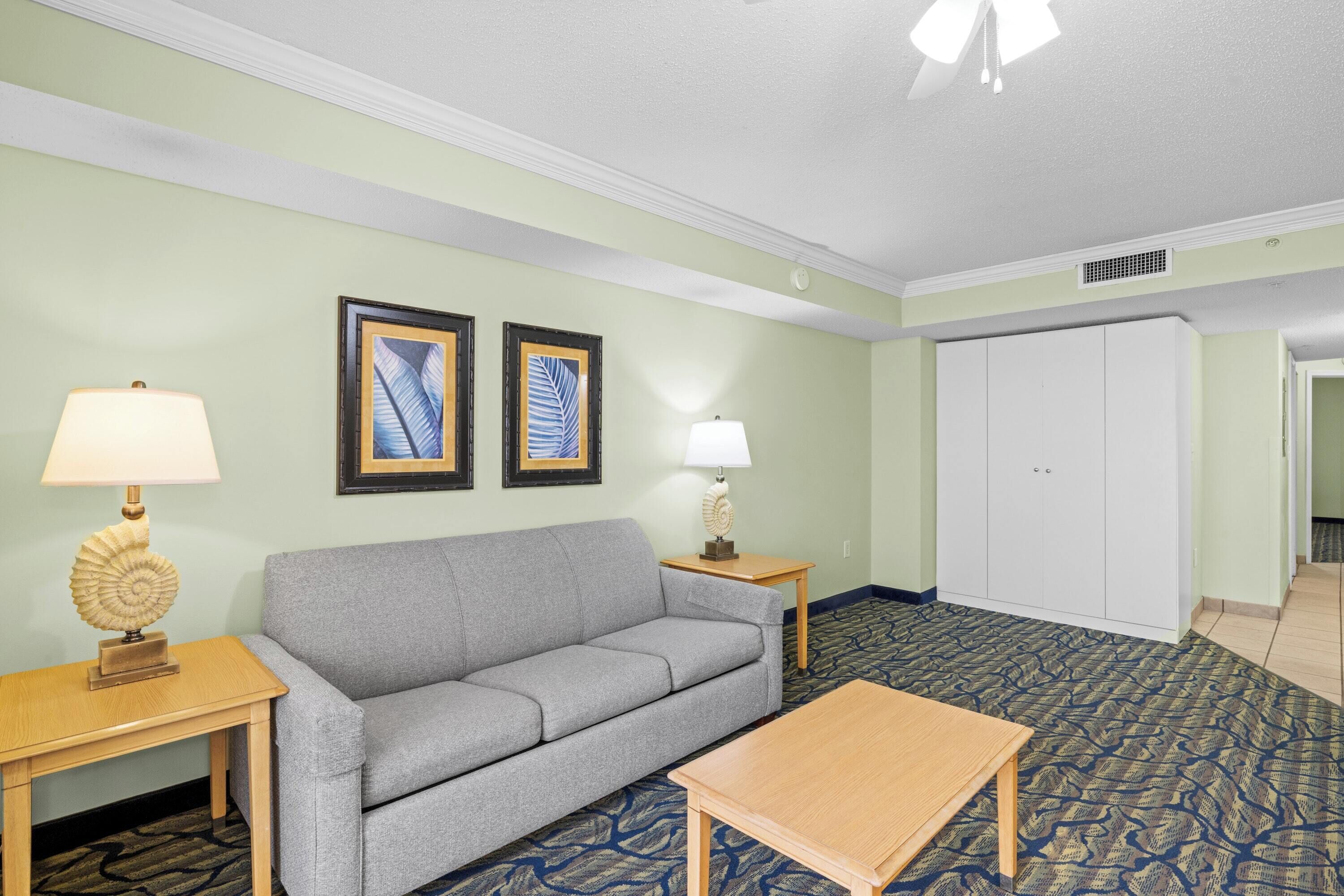 A hotel room with a grey sofa, wooden side tables, a wooden coffee table, a patterned carpet, and a white wardrobe.