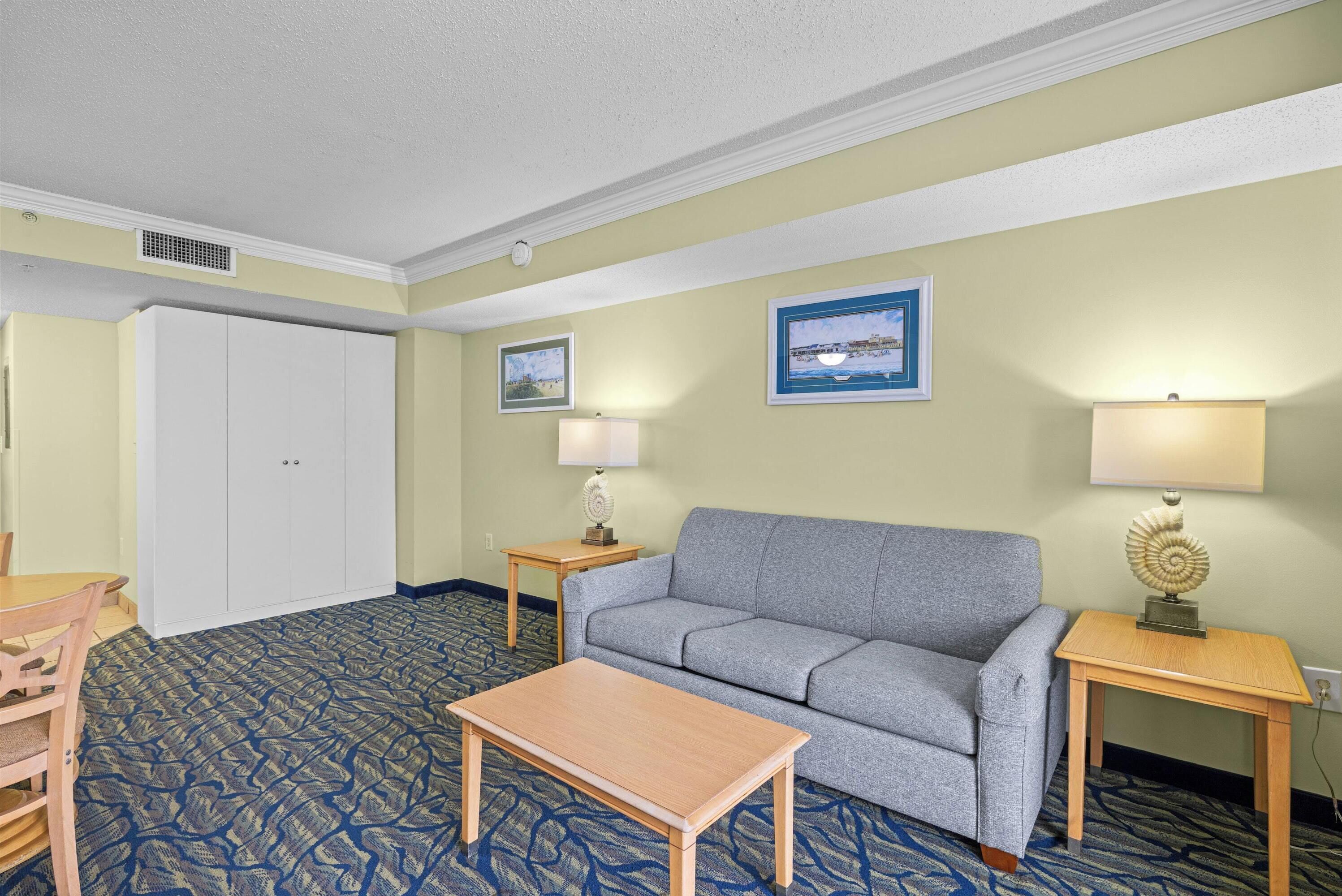 A hotel room with a grey sofa, a wooden coffee table, a dining table with chairs, two bedside tables with lamps, and a door.
