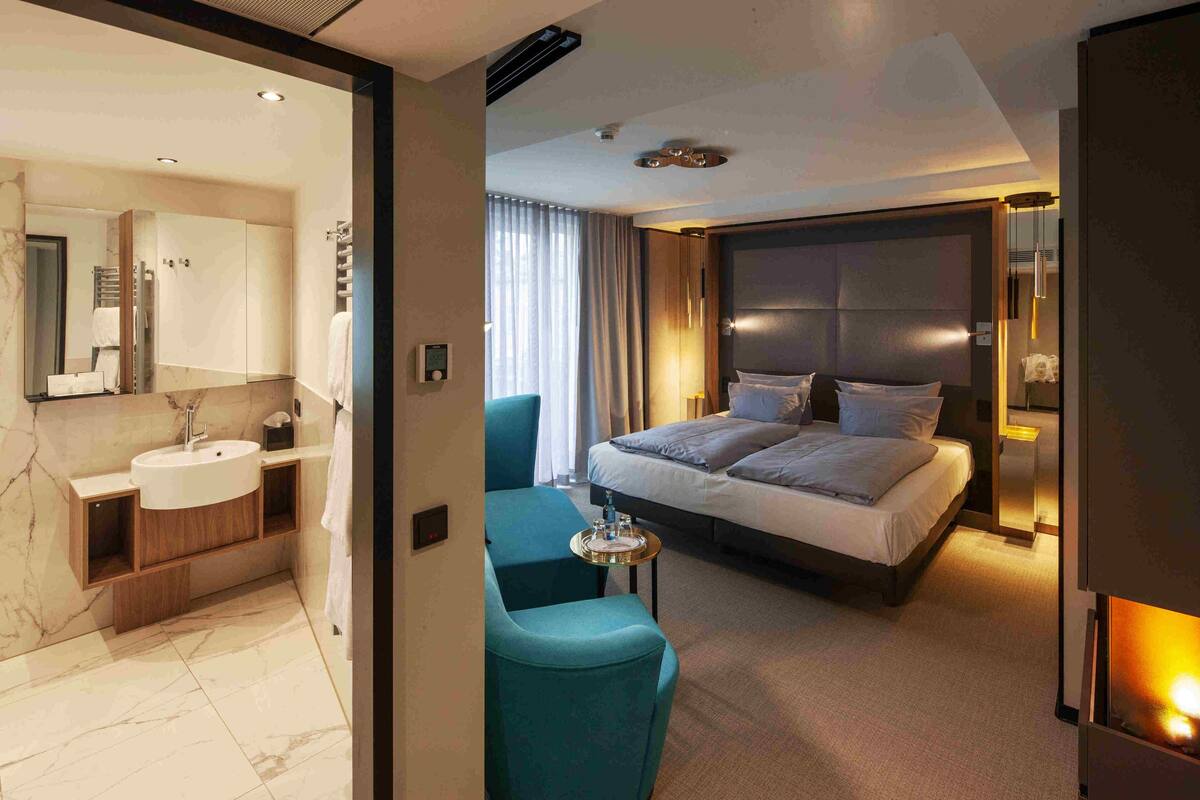 Une chambre d’hôtel moderne dotée d’une salle de bain en marbre, d’un grand lit avec une tête de lit et d’un coin salon avec une petite table.