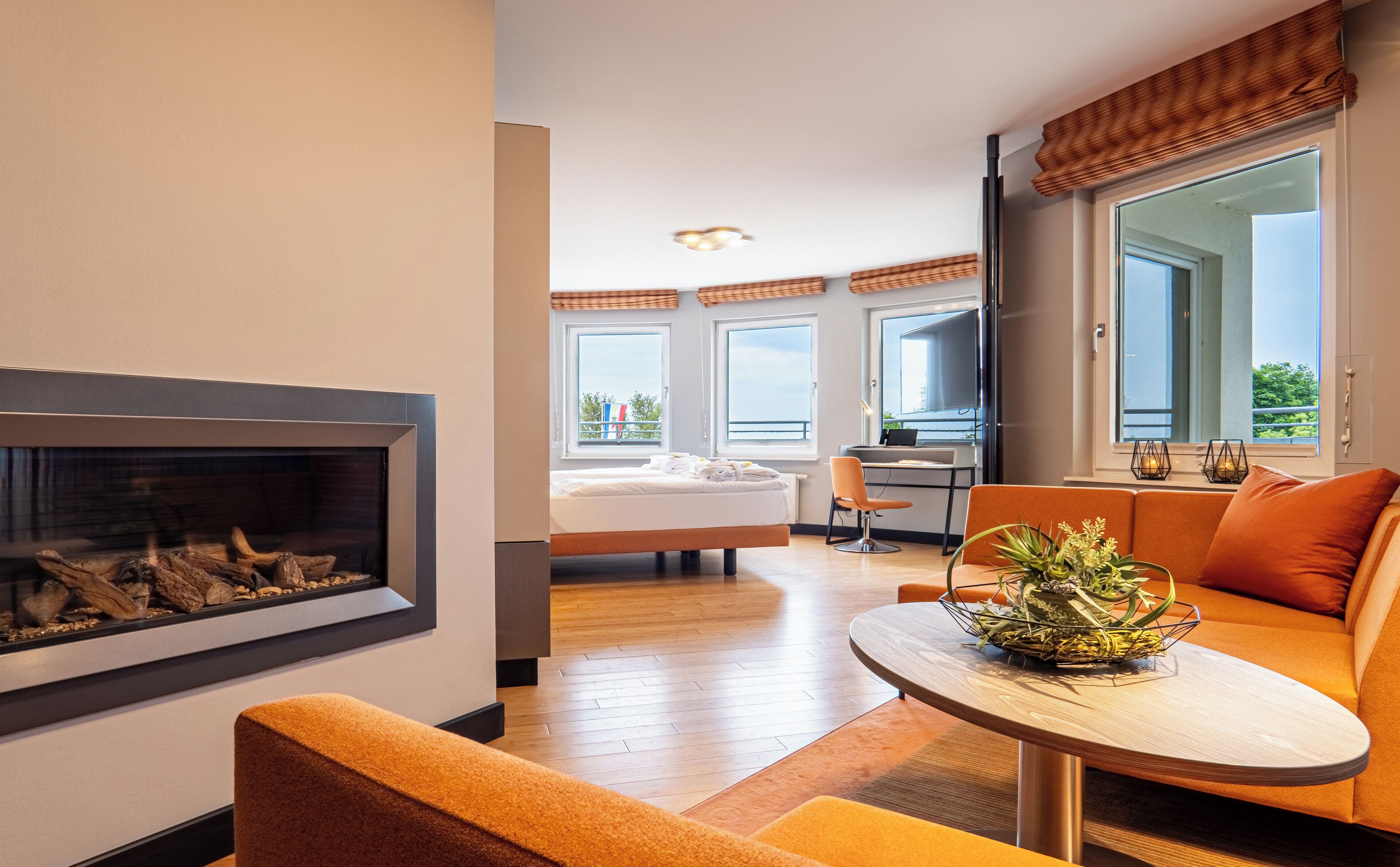 A modern hotel room with a fireplace, a dining area with a table and orange sofa, a bed with a view, and large windows.