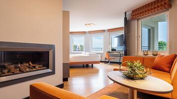 A modern hotel room with a fireplace, a dining area with a table and orange sofa, a bed with a view, and large windows.
