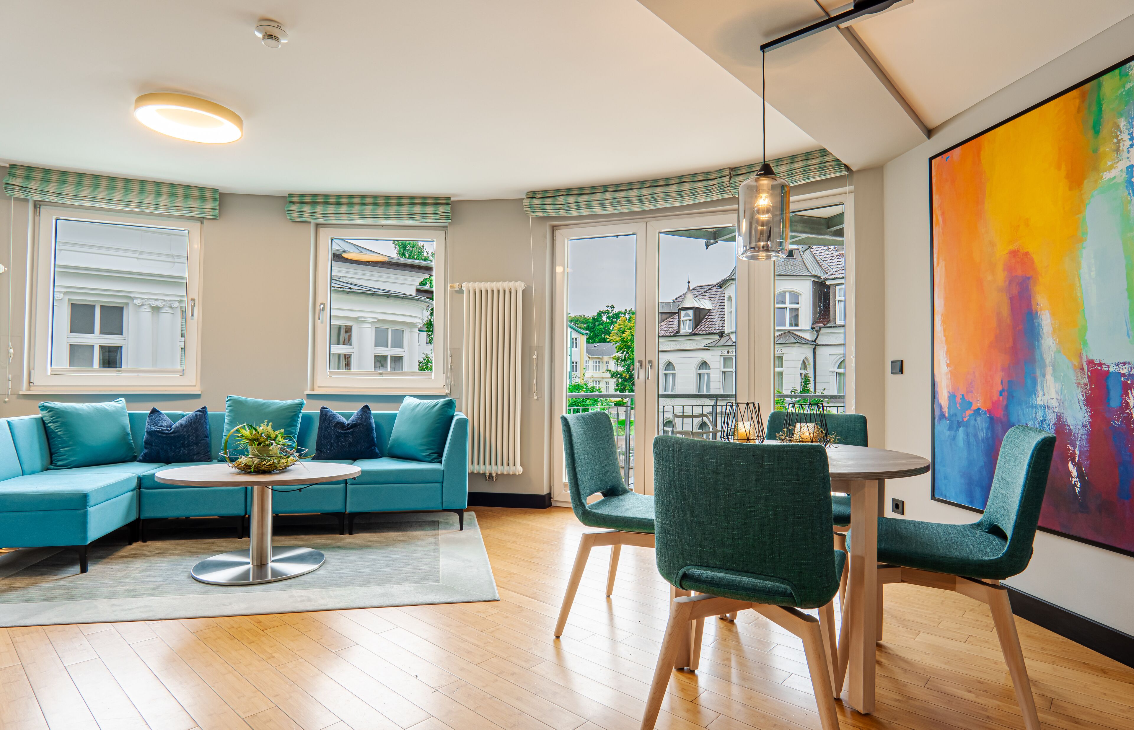A modern living room with a blue sofa, a round table, and green chairs. There is a large window with a view of buildings outside.