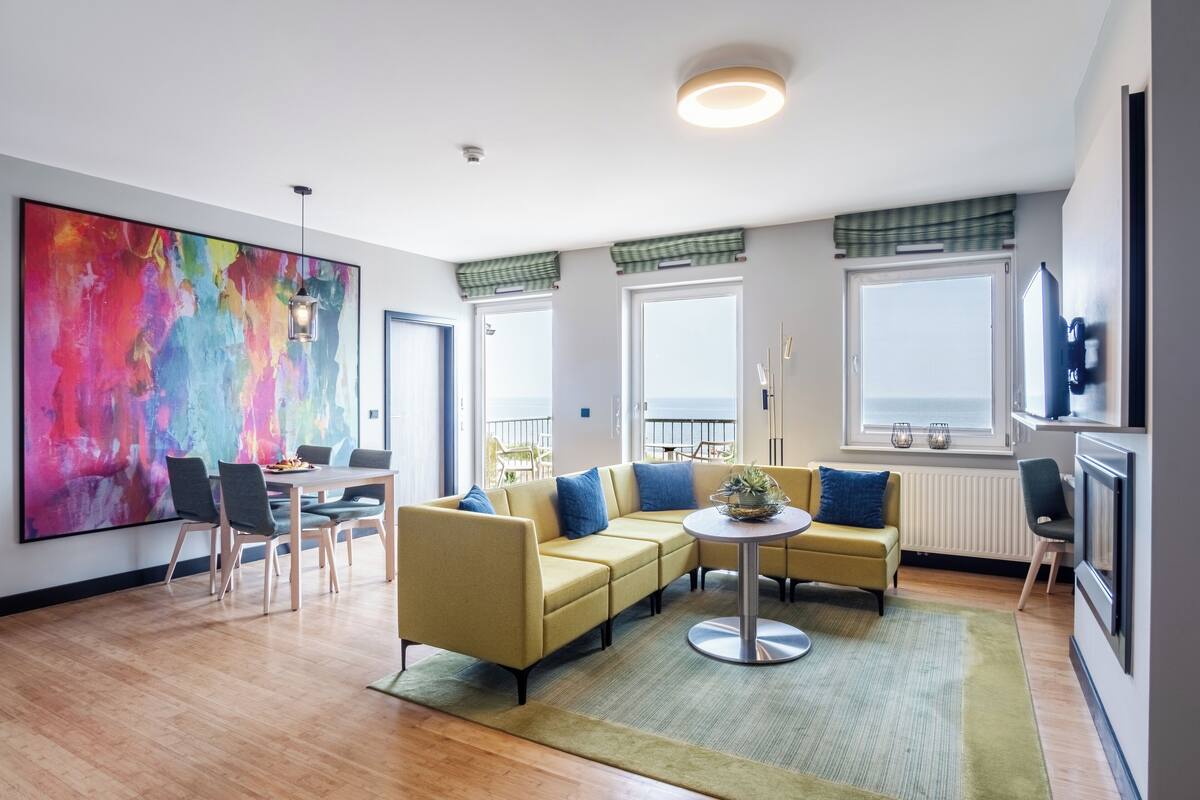 Un salon moderne avec un canapé d’angle, une table à manger et des chaises. Un grand tableau abstrait orne le mur, ainsi qu’une lampe sur pied et une petite table ronde sur laquelle est posée une plante.