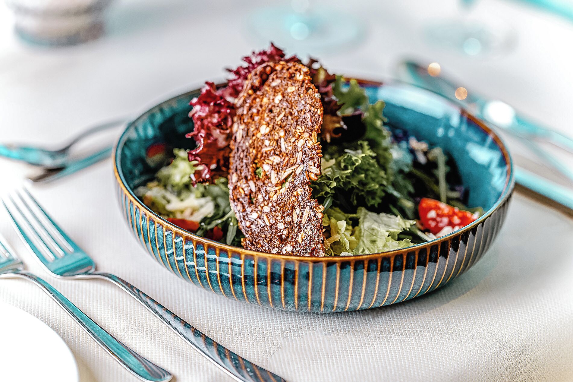A bowl of salad with a slice of bread on top, placed on a table with a blue and white striped bowl.