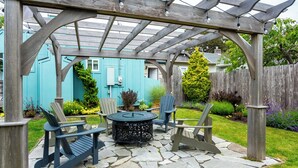Un área de patio techada con sillas Adirondack y una mesa redonda, rodeada por una pérgola de madera y una casa azul al fondo.