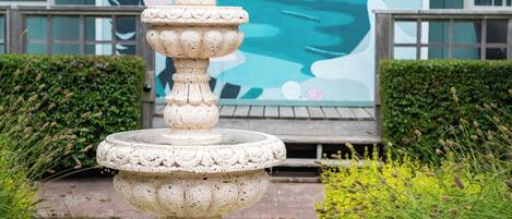 A stone fountain with a tiered design in a garden setting, surrounded by greenery.